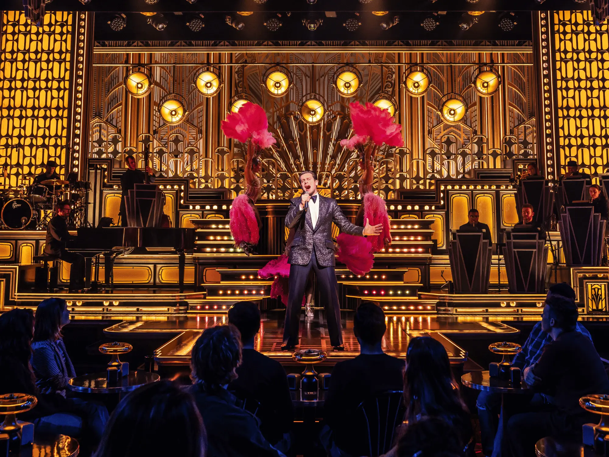 A singer flanked by pink feather fans performing on a glittering gold art deco Broadway stage